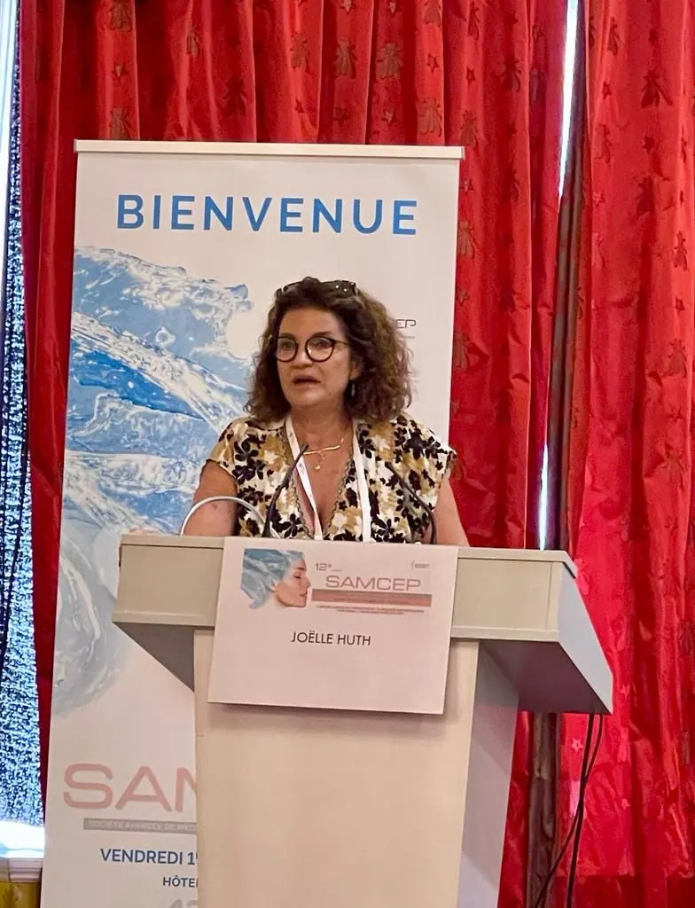 Le Dr Joëlle Huth au pupitre d’un congrès de médecine esthétique, devant une bannière « Bienvenue »
